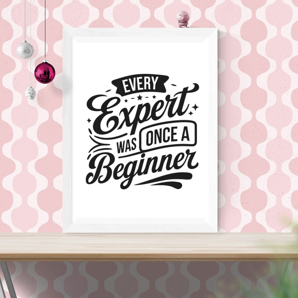 Black stamp-style seal print reading Every Expert Was Once a Beginner in a white frame on a pink patterned wall.