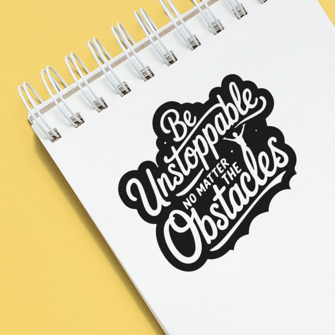 Black-and-white stamp seal reading Be Unstoppable No Matter the Obstacles on a white spiral notebook over yellow