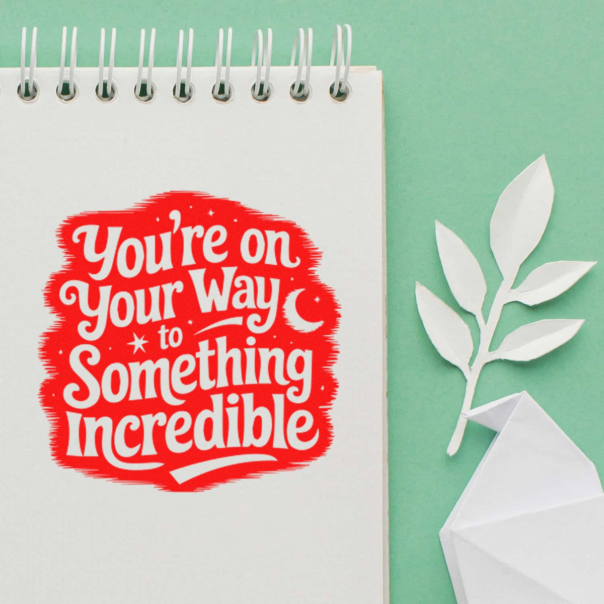 Red motivational stamp on white spiral notebook reads Youre on your way to something incredible with white paper leaf seal on mint background.