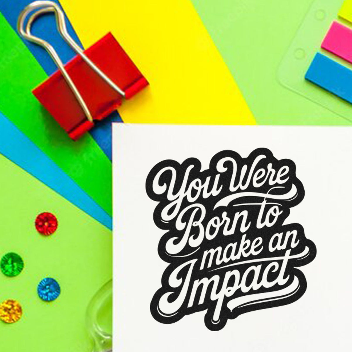 Bright green, yellow, and blue stationery with a red binder clip and black script stamp seal reading You Were Born to make an Impact