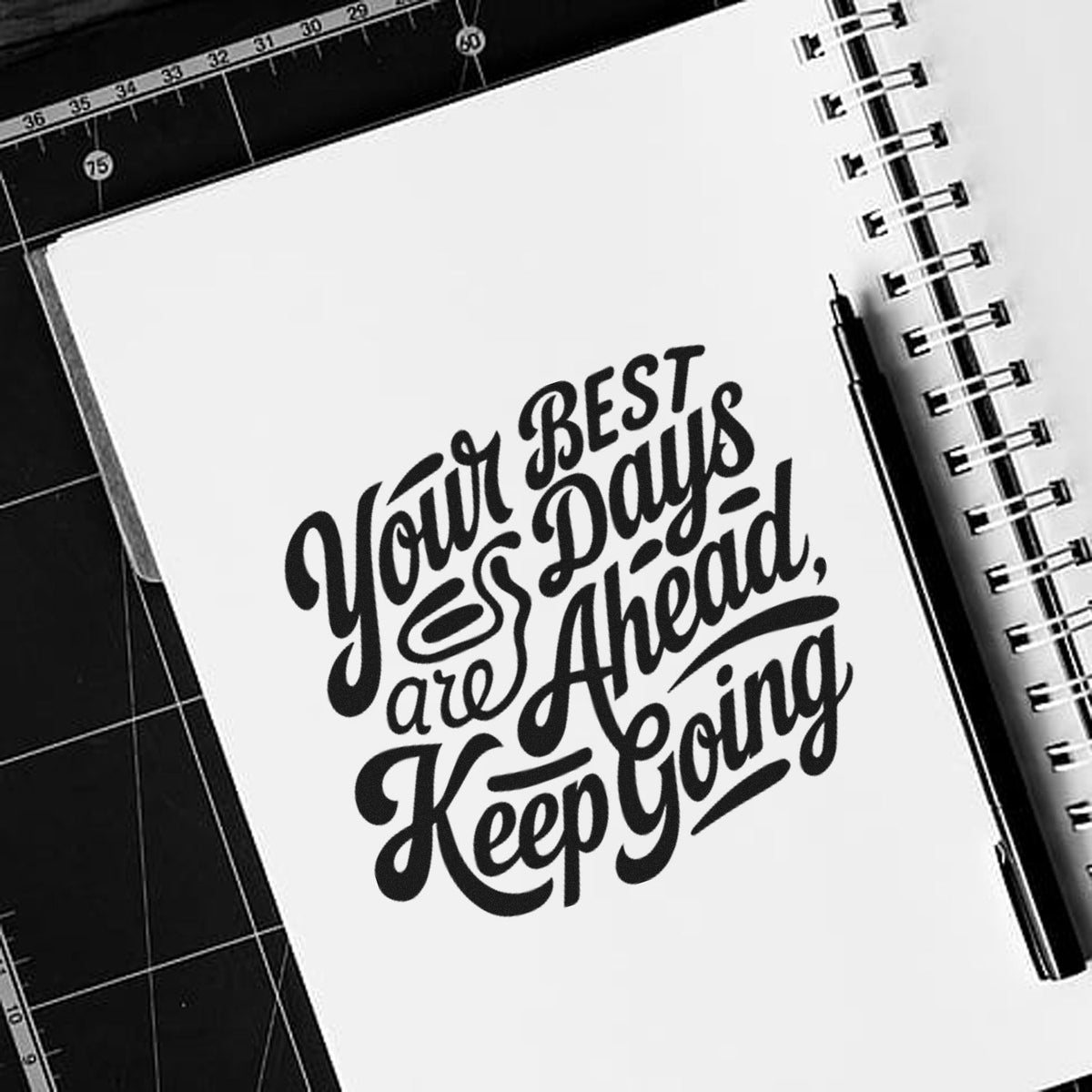 Black quote stamp/seal on a white spiral notebook, with pen on cutting mat: Your best days are ahead, keep going.