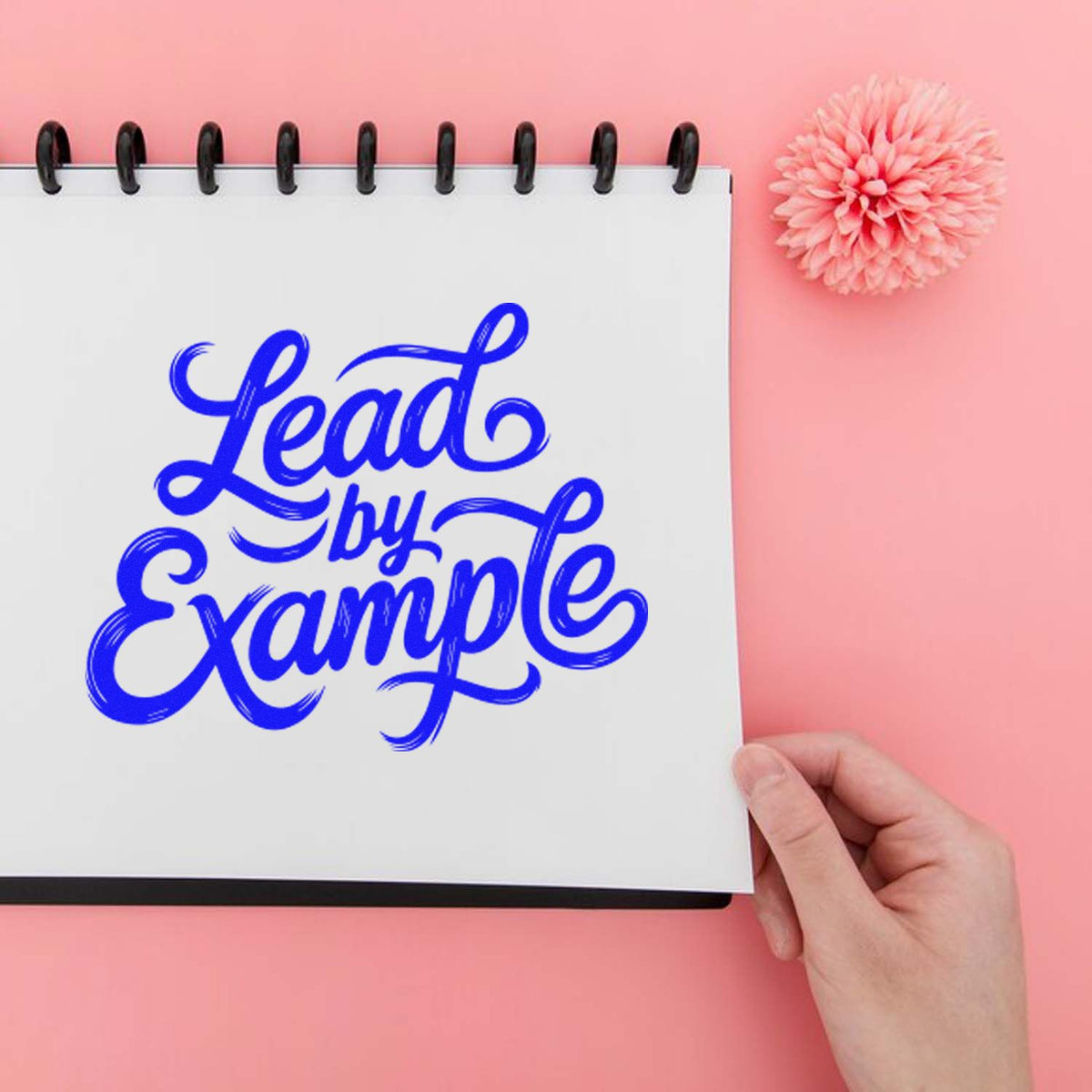 Blue Lead by Example stamp/seal design on a white spiral notebook, with a pink flower on a pastel pink background.