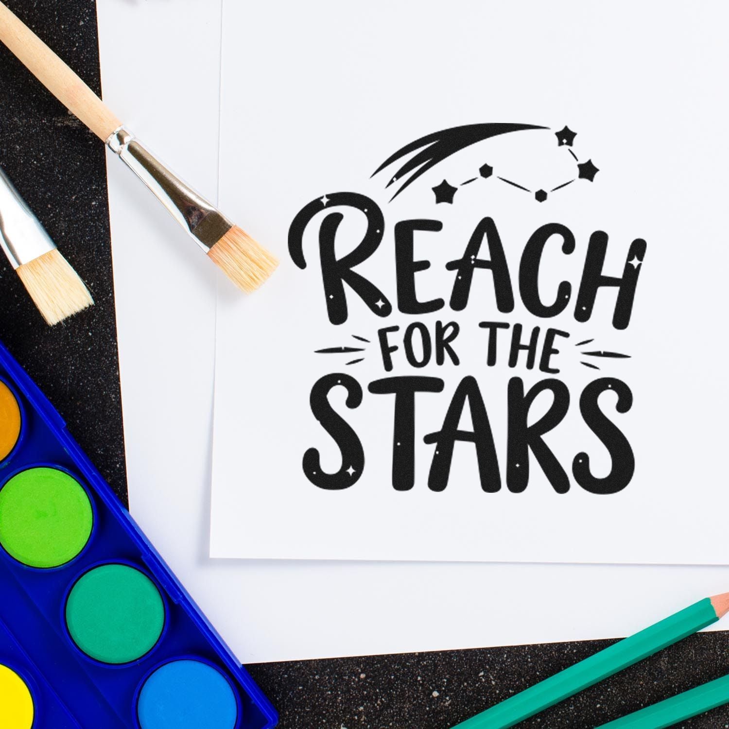 Black Reach for the Stars stamp design on white paper, surrounded by paintbrushes, bright watercolor pans, and pencils.