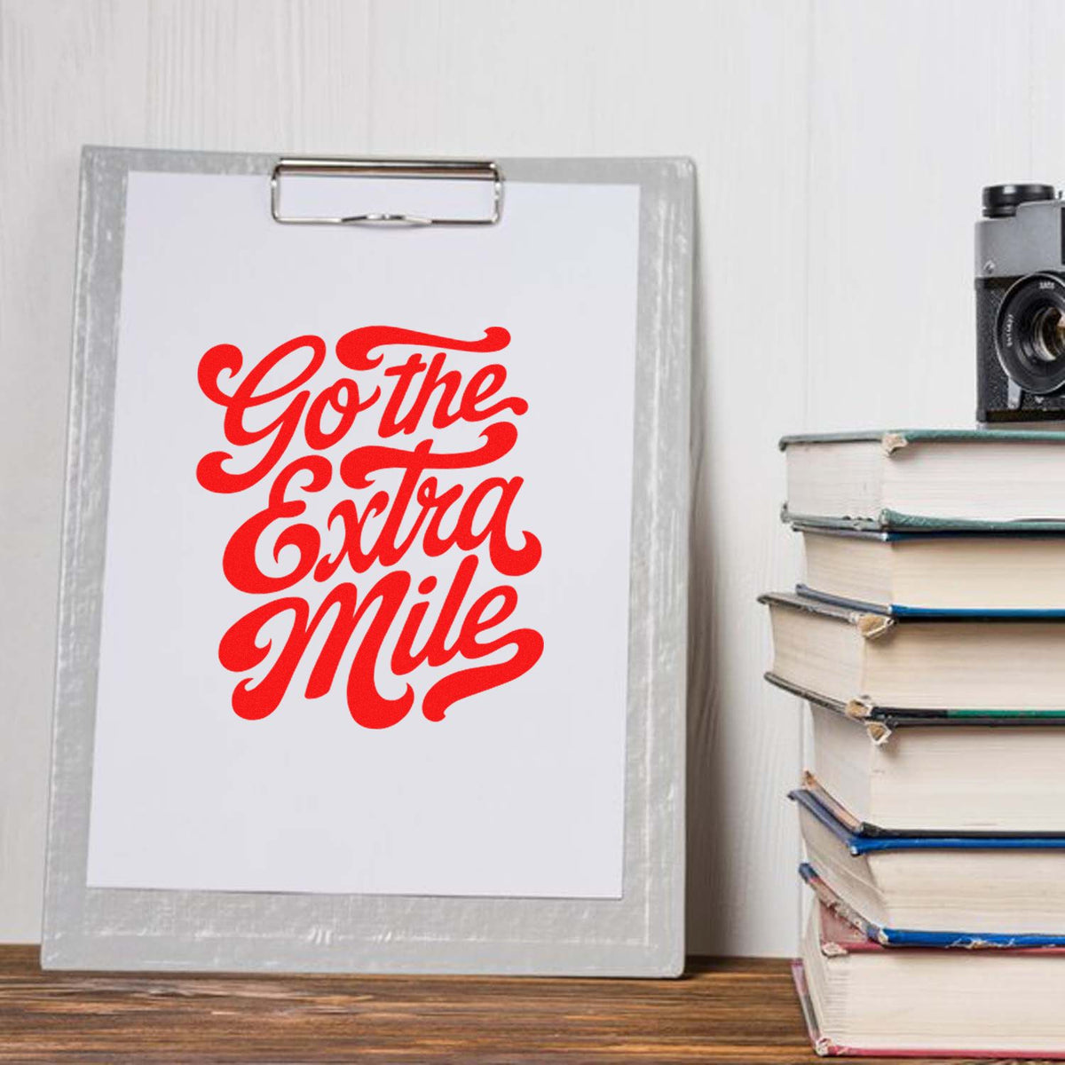 Red Go the Extra Mile custom stamp and seal design on white paper clipped to a silver clipboard, beside stacked books.