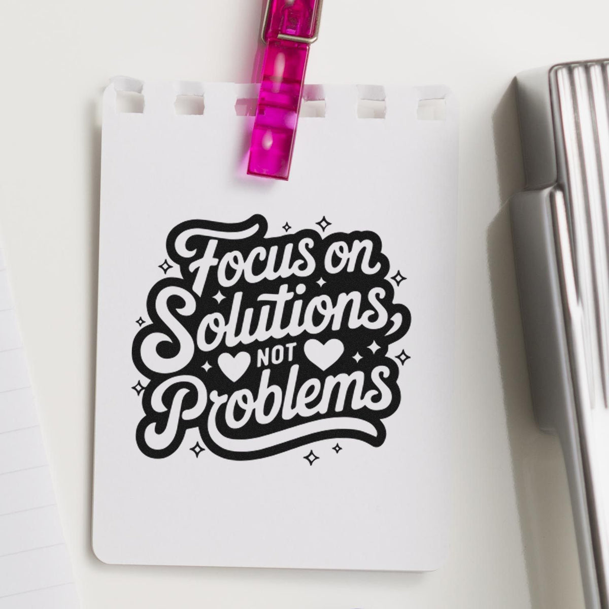 Black script stamp on white notepad, clipped with pink seal, reading Focus on Solutions, Not Problems with hearts and sparkles.