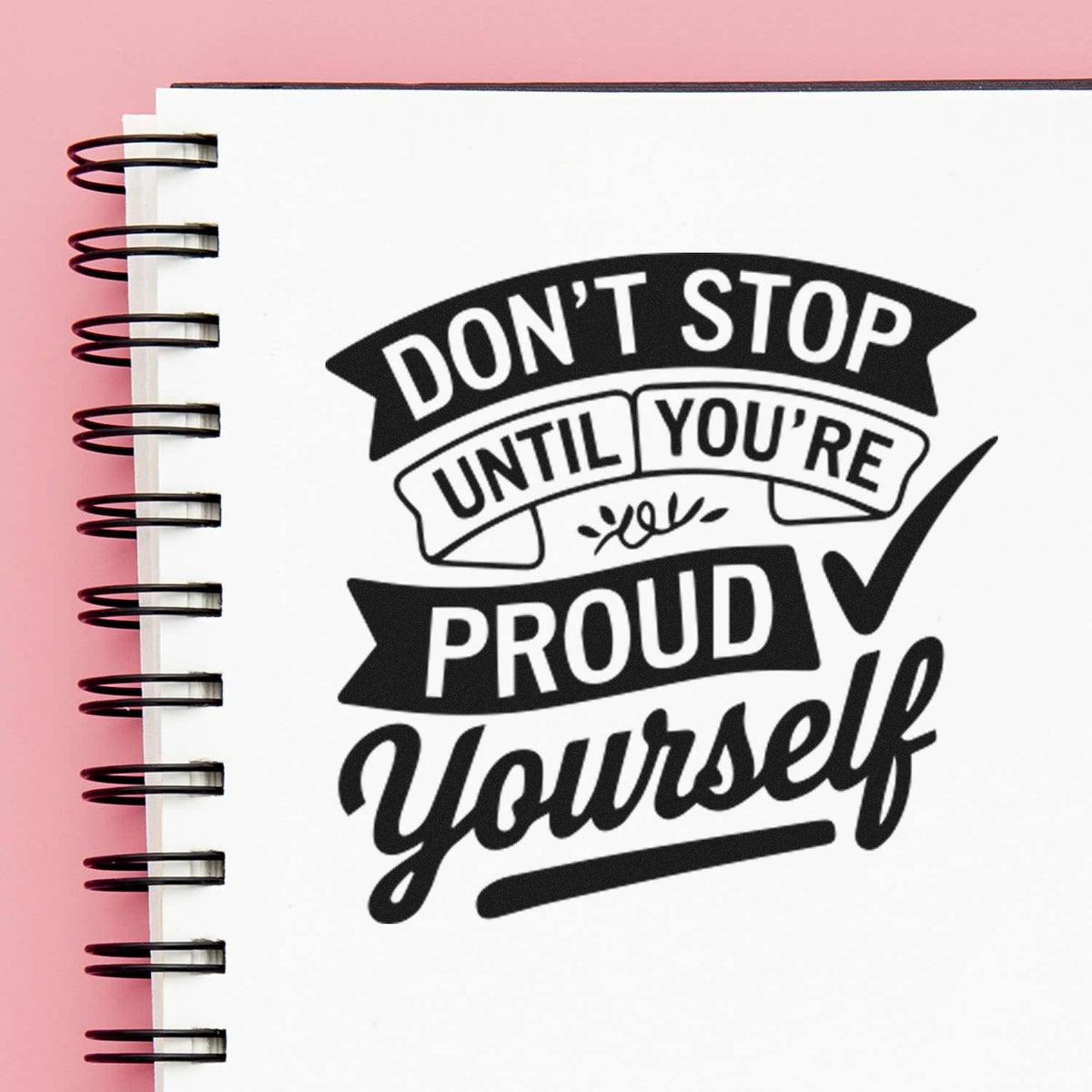 Black motivational stamp and seal reading Dont stop until youre proud yourself on white spiral notebook with pink background.