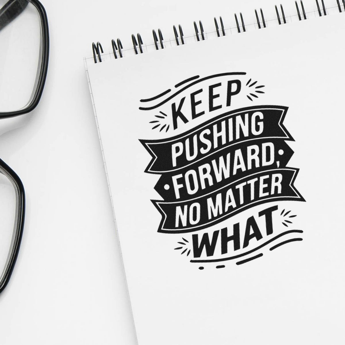 Black motivational stamp on a white spiral notebook, reading Keep pushing forward, no matter what, with black glasses nearby.