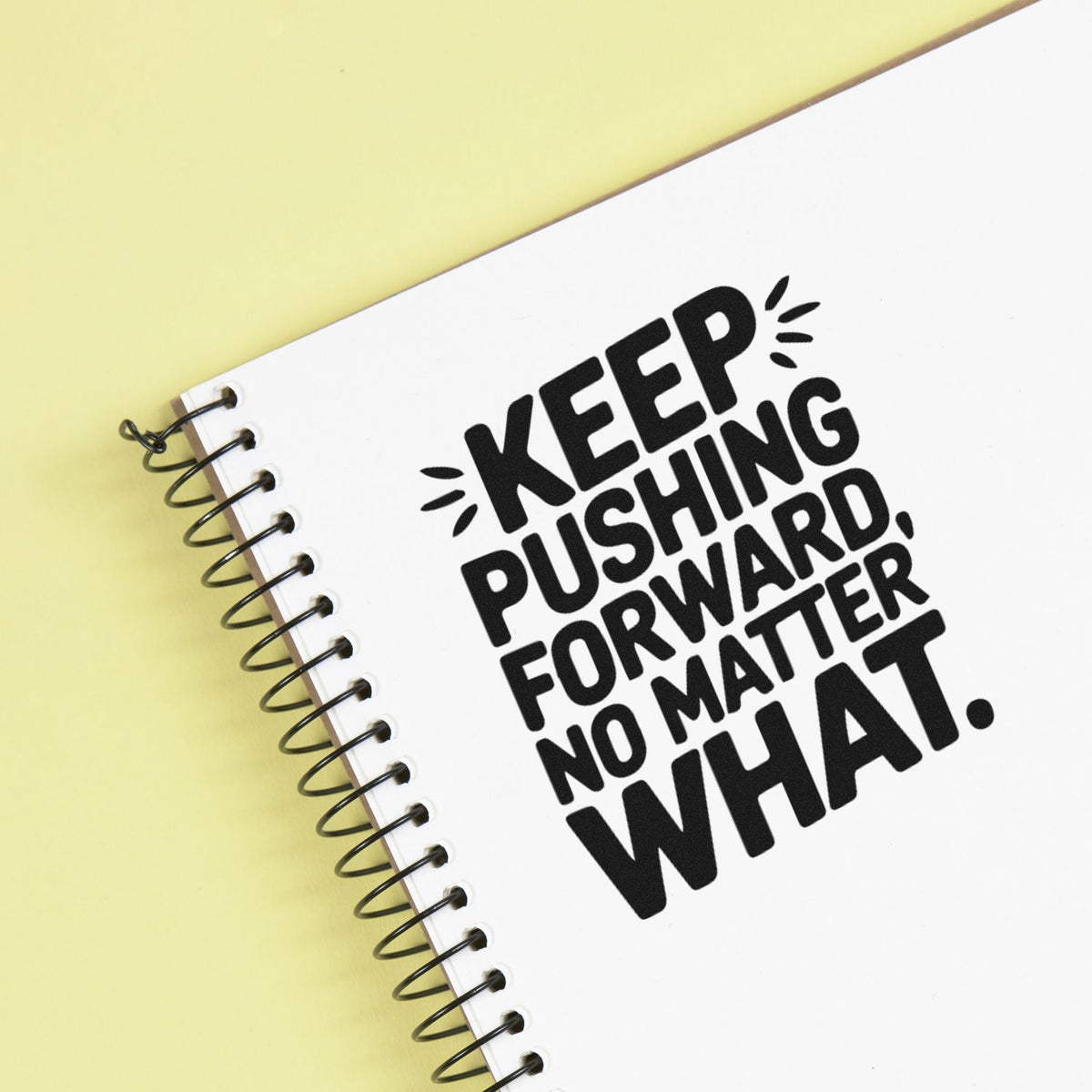 Black rubber stamp design on white spiral notebook over pale yellow background, with bold Keep pushing forward, no matter what seal.