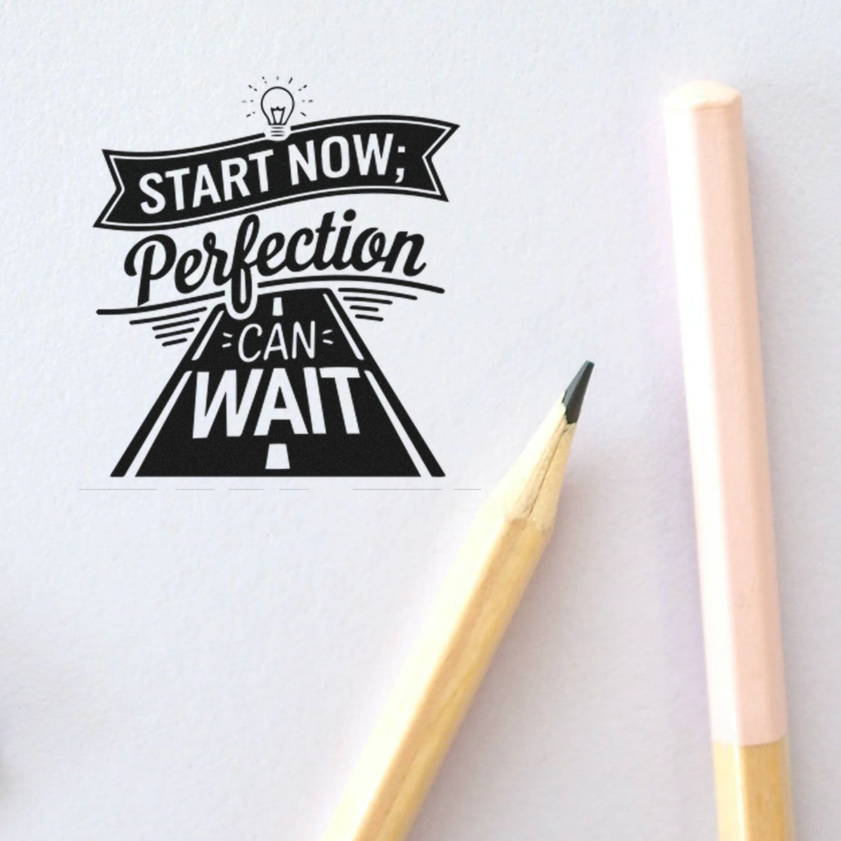 Black motivational stamp seal reading Start Now; Perfection Can Wait on white paper beside a pencil and pink-handled brush.