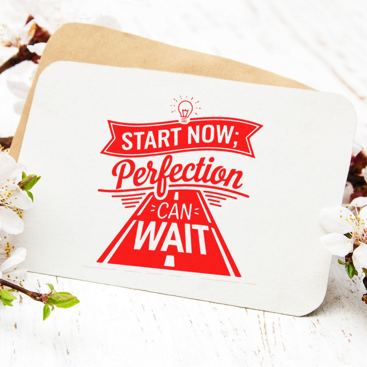 Red rubber stamp seal on white card reading Start Now; Perfection Can Wait with a lightbulb icon, on tan envelope with white flowers.