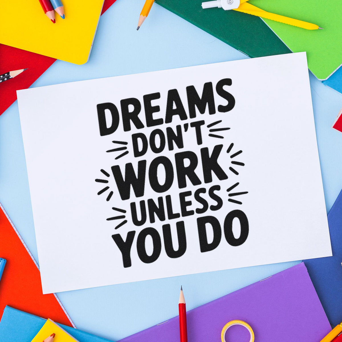 Black motivational rubber stamp design reading "Dreams don't work unless you do" on white paper, framed by colorful pencils and notebooks.