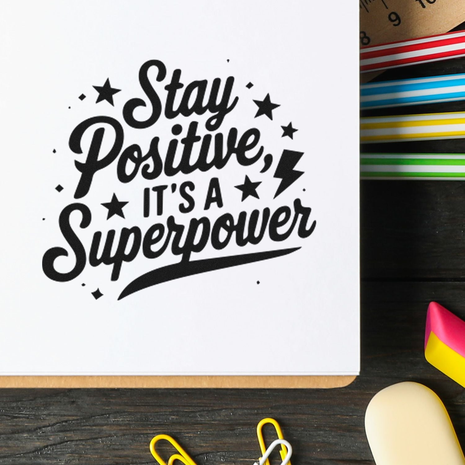 Black motivational stamp reading Stay Positive, Its A Superpower on white paper, with colorful seal strips and office supplies.
