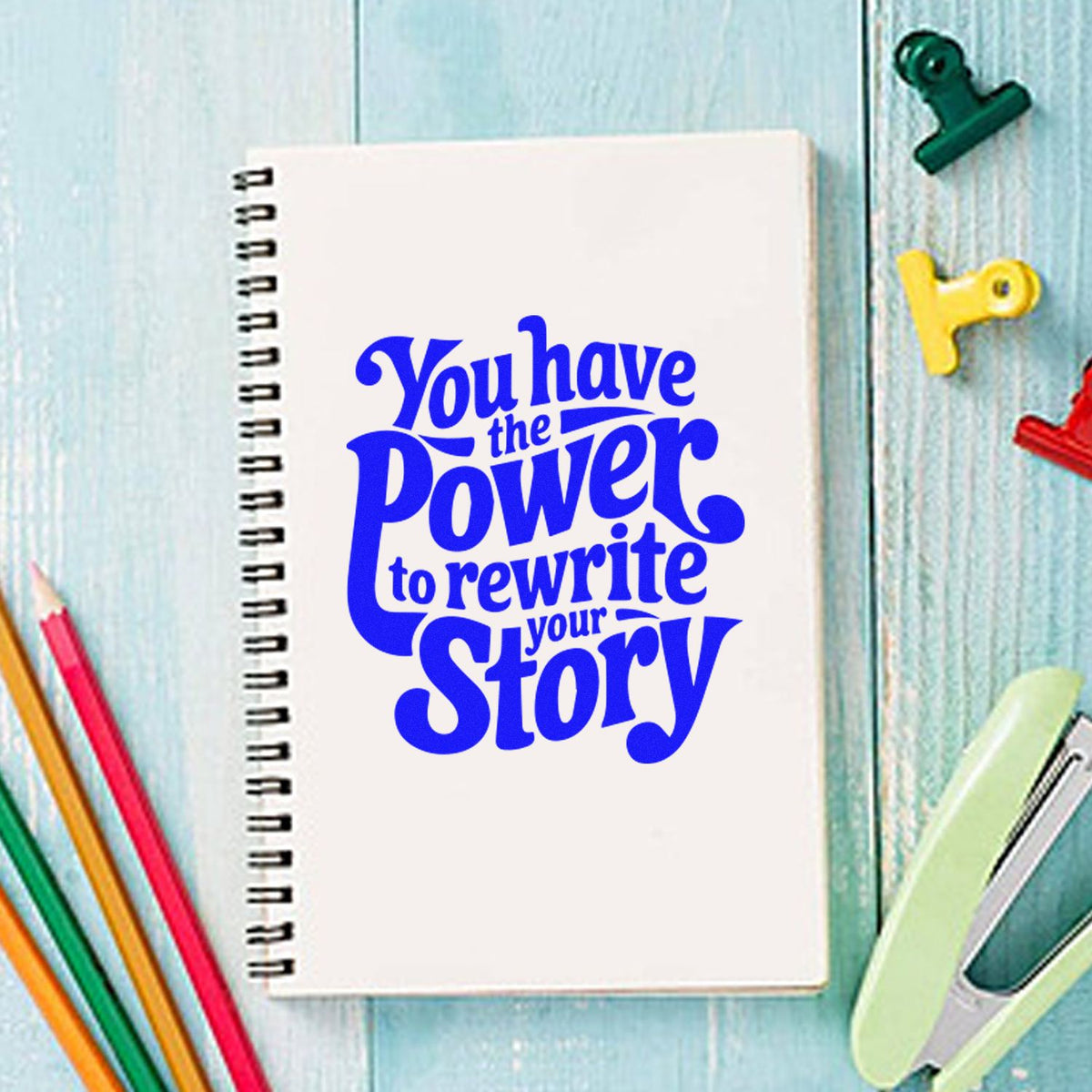 White spiral notebook with bold blue stamp design, surrounded by colorful stamp and seal products on a light blue desk.