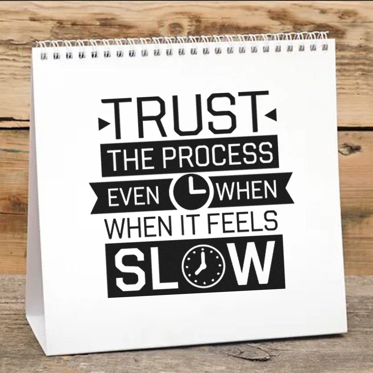 Black stamp and seal design on a white spiral-bound desk calendar, reading Trust the process even when it feels slow.