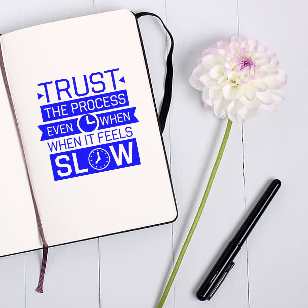 White notebook with a blue stamp/seal design reading Trust the process, beside a black pen and white flower.