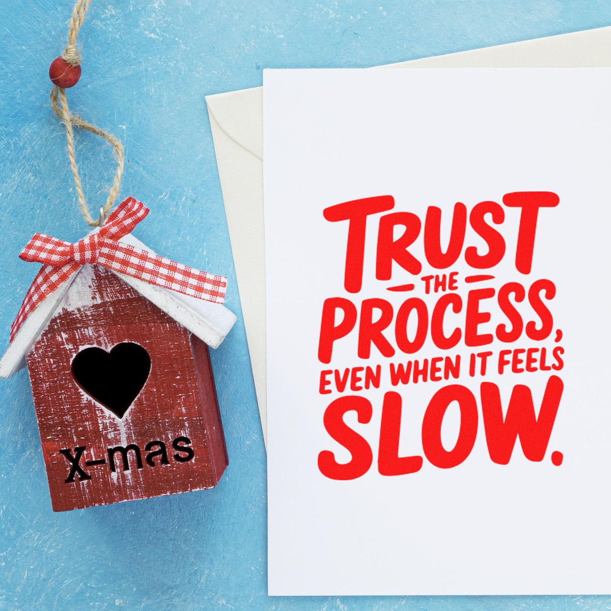 Red house-shaped Christmas stamp seal with heart cutout beside a card saying Trust the process, even when it feels slow.