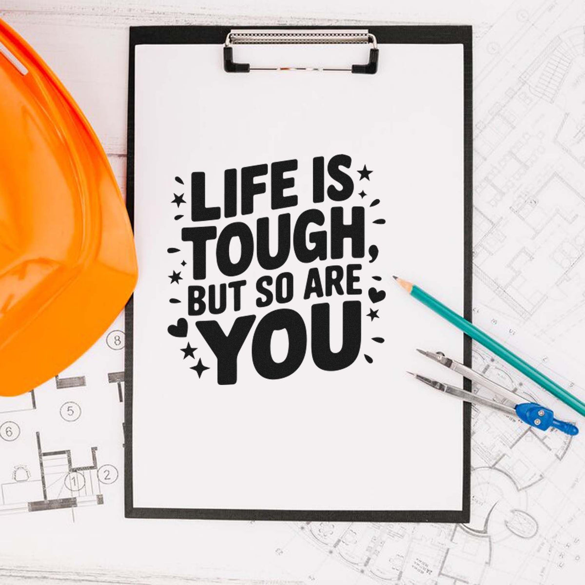 Custom stamp on clipboard reads Life is tough, but so are you, beside orange hard hat, pencils, and blueprints.