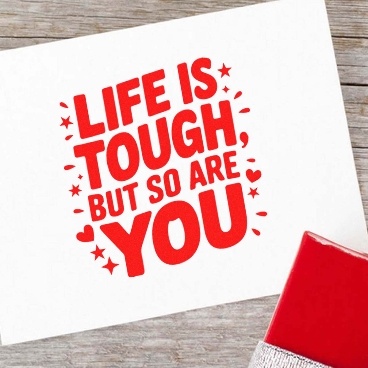 Red rubber stamp impression reading Life is tough but so are you on white paper, with a red stamp block on wood.