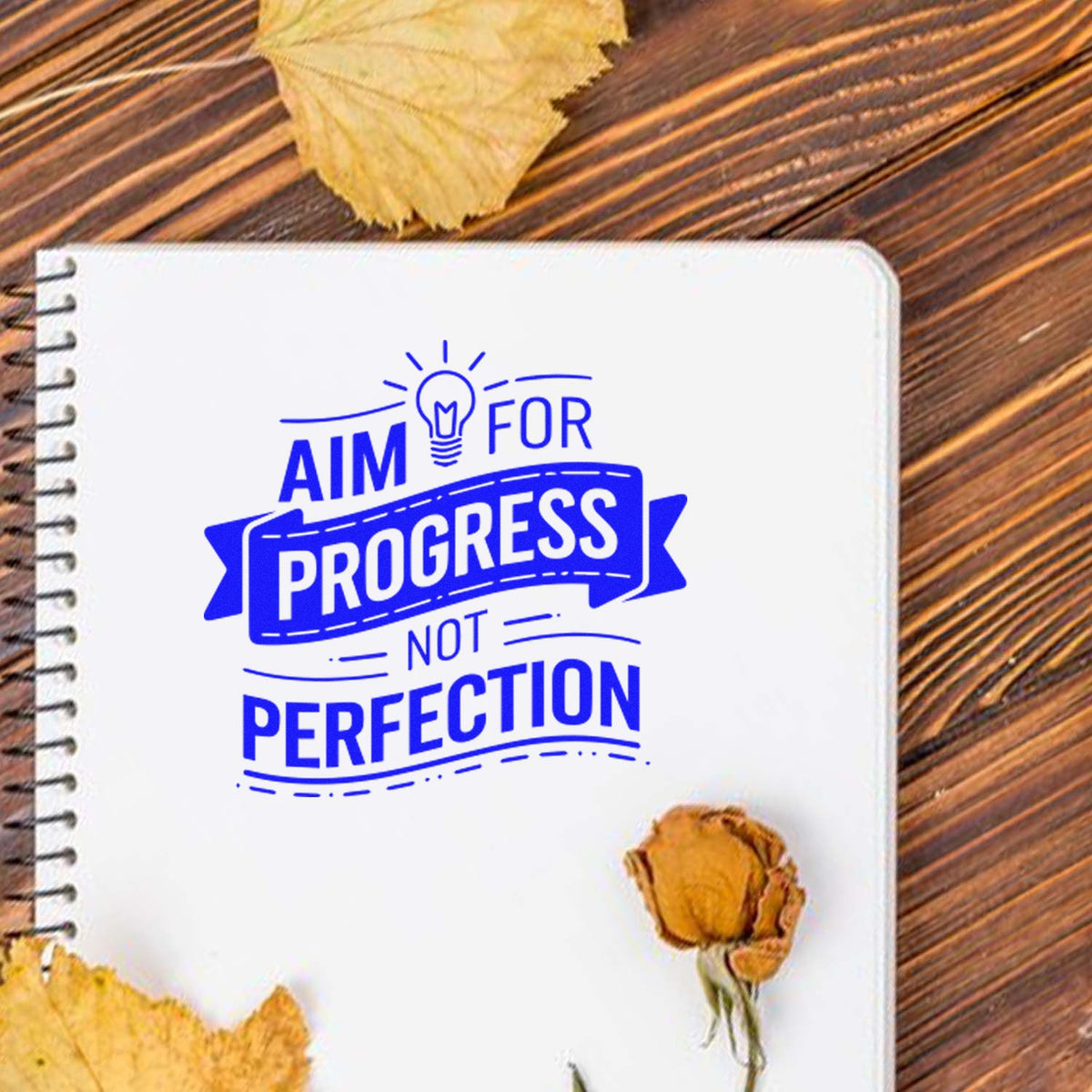 Blue motivational stamp seal on white notebook page reading Aim for progress, not perfection, on a wood table with dried leaves.