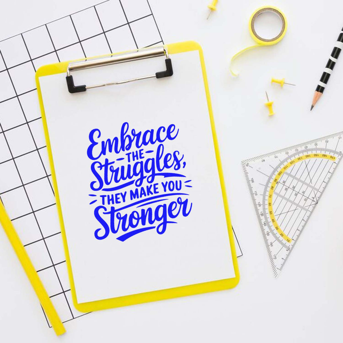 Blue motivational stamp/seal on a yellow clipboard, with rulers and drafting tools on a white background.