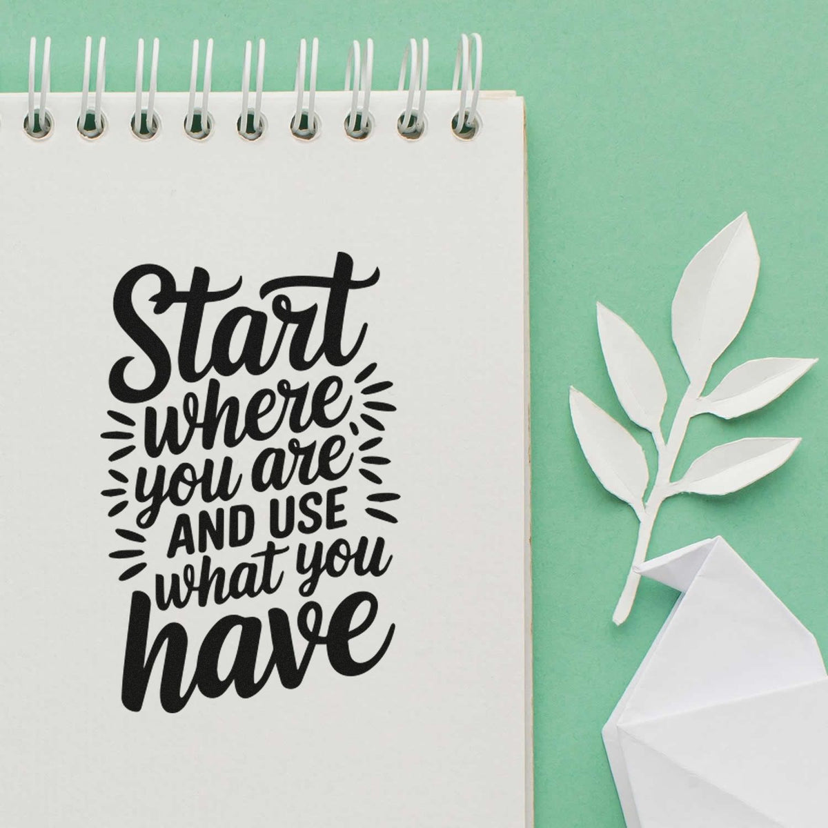 Black quote stamp on white spiral notebook: Start where you are and use what you have, with white paper leaves on mint green.