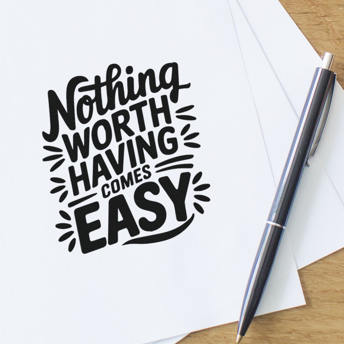 Black custom stamp impression reading Nothing Worth Having Comes Easy on white paper, with a sleek black pen on a wood desk.