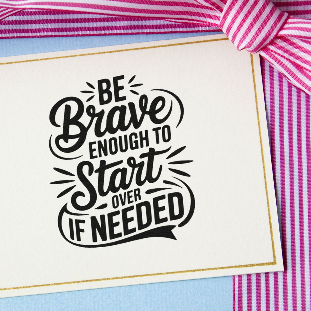 Black stamp/seal with motivational text on white card, gold border, pink striped ribbon, and blue background: Be Brave Enough to Start Over If Needed