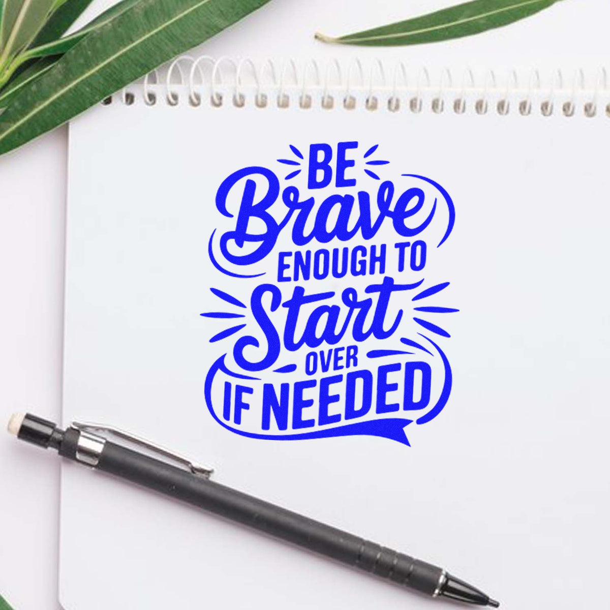 Blue custom stamp seal reading Be Brave Enough to Start Over If Needed on a white notebook, with pen and green leaves.