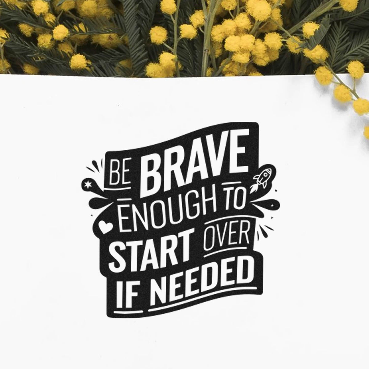 Black decorative stamp seal with white text, Be brave enough to start over if needed, on white paper with yellow flowers.