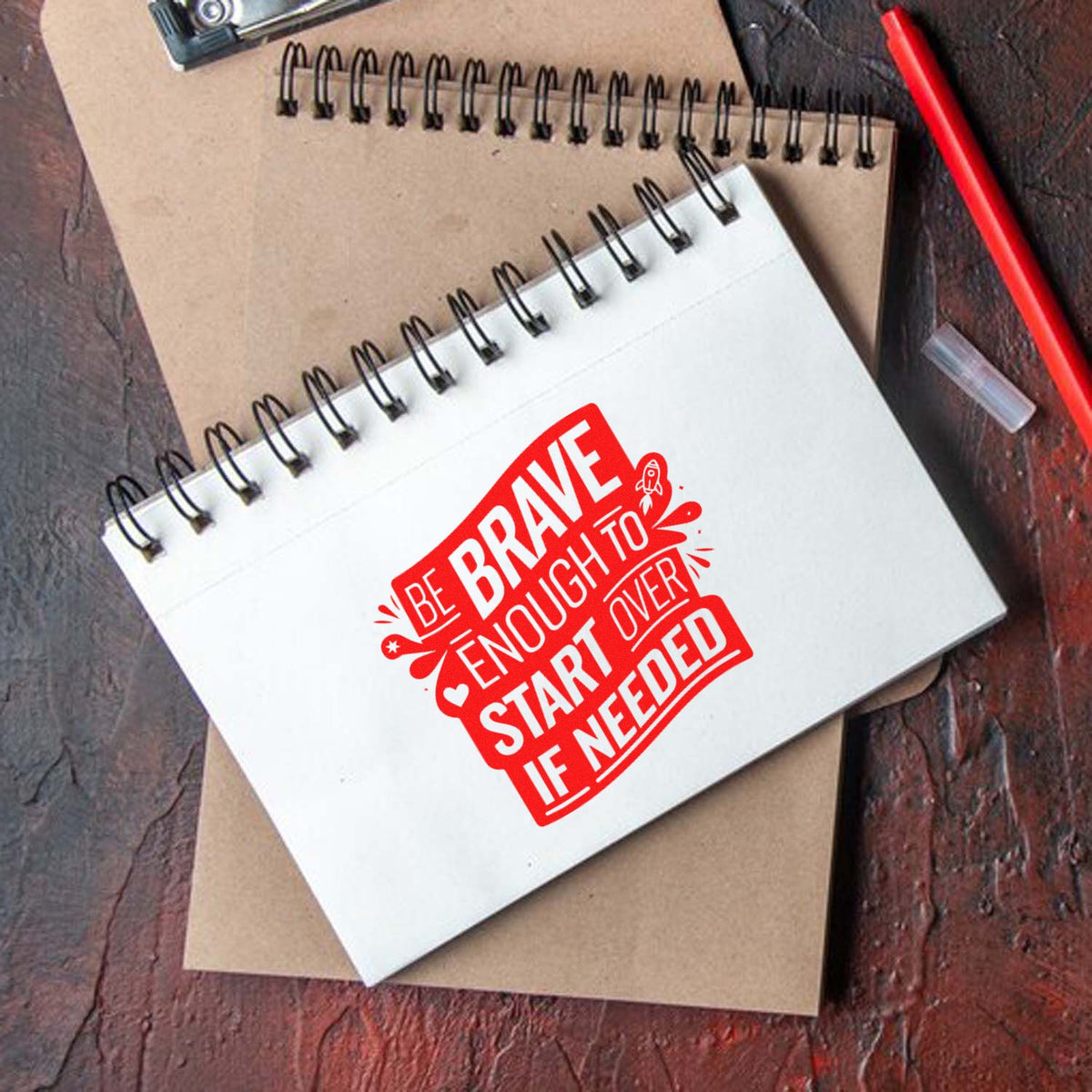 White spiral notebook with a red custom stamp seal reading Be brave enough to start if needed, on brown notebook, beside a red pen.