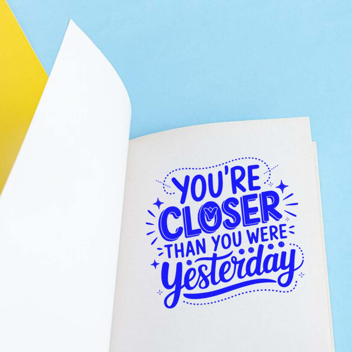 Blue stamp-style seal on white paper reading Youre Closer Than You Were Yesterday against a light blue background.