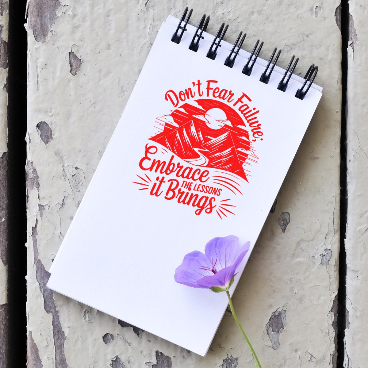 White spiral notebook with a red motivational stamp/seal design and a purple flower on weathered wood.
