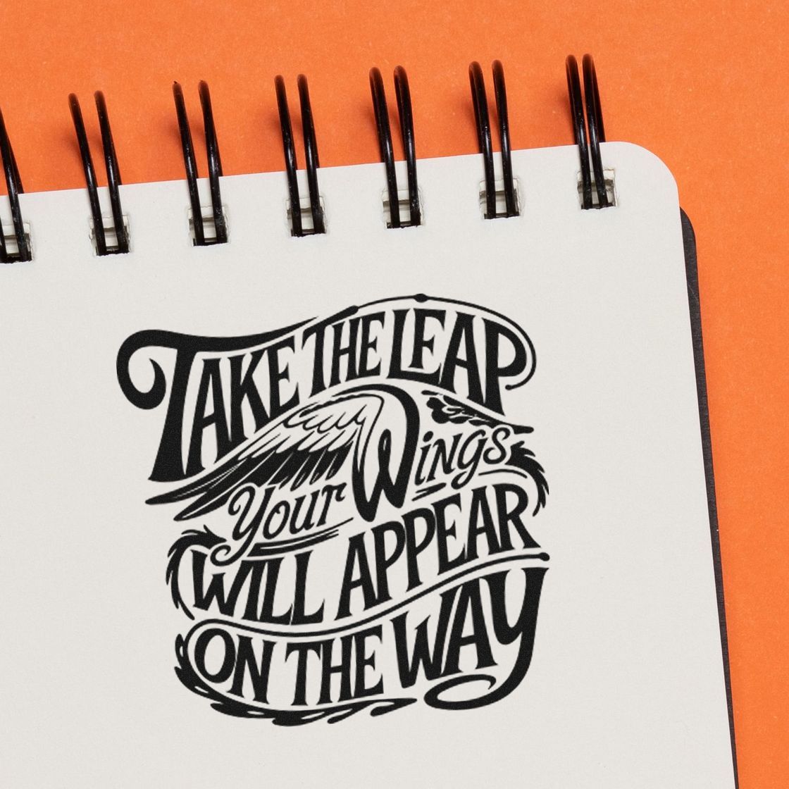 Black decorative stamp imprint with Take the Leap quote and wing design on a white notebook page, orange background.