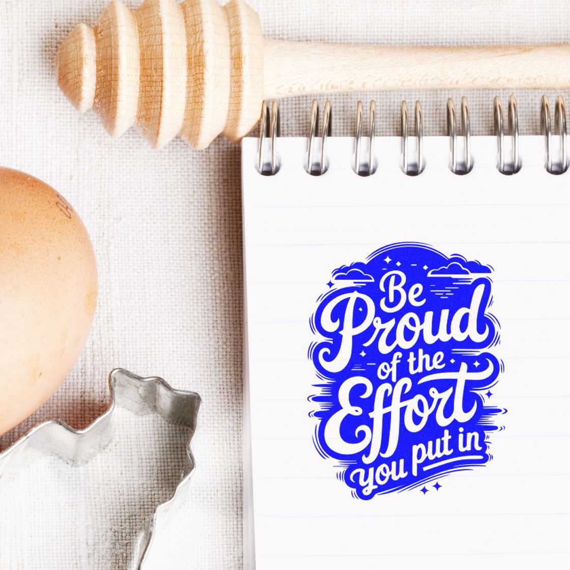 Blue motivational rubber stamp seal on a lined notebook, beside a wooden honey dipper, egg, and metal cutter.