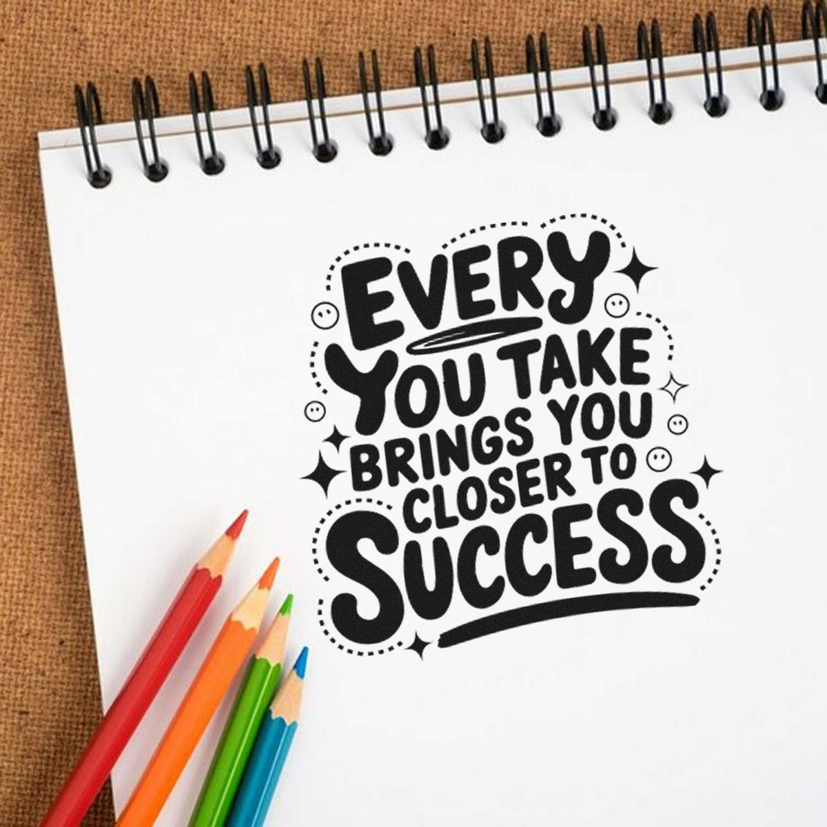 Black motivational stamp/seal on white spiral notebook with colored pencils, reading Every you take brings you closer to success.