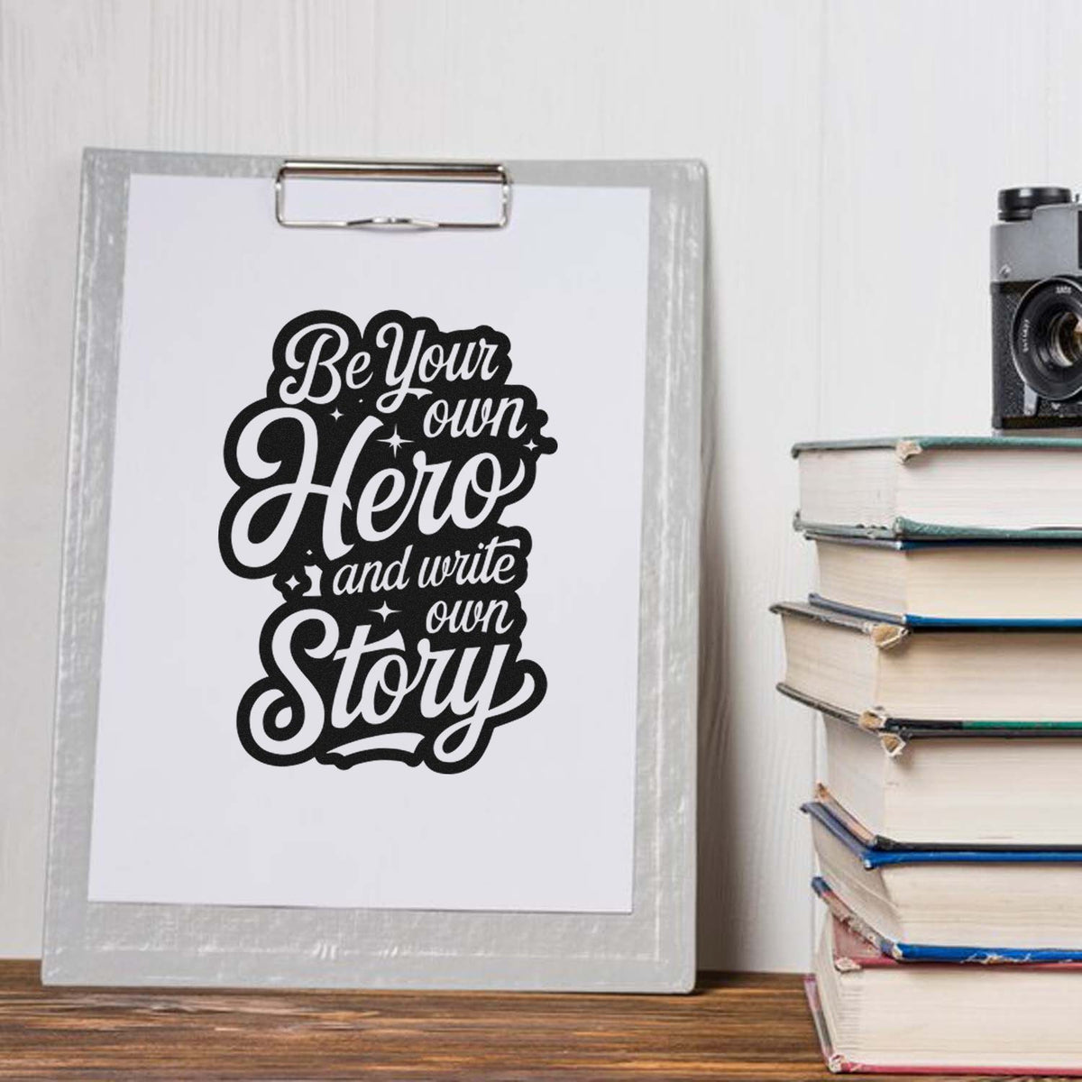 Black custom stamp and seal design with Be Your Own Hero and write own Story on a white clipboard beside stacked books.
