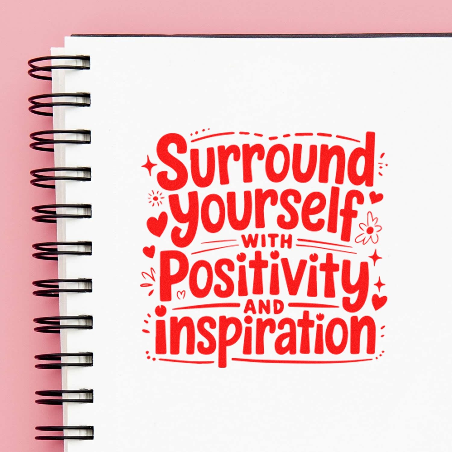 Red stamp/seal design on a white spiral notebook cover reading Surround yourself with positivity and inspiration on a pink background.