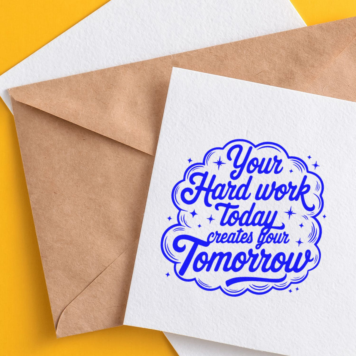 Blue rubber stamp seal with motivational text on a white card beside kraft envelopes on a yellow background.