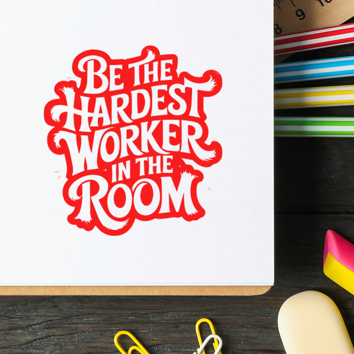 Red stamp seal reading Be the Hardest Worker in the Room on white paper, with colorful pens nearby.