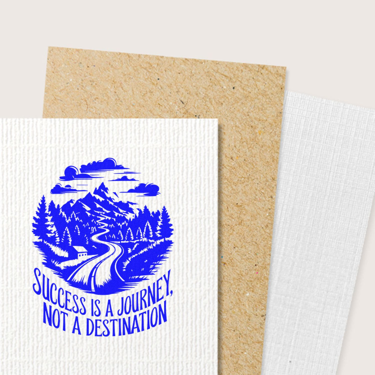 Blue rubber stamp seal on white textured paper, featuring mountain road, pine trees, and Success is a journey, not a destination.