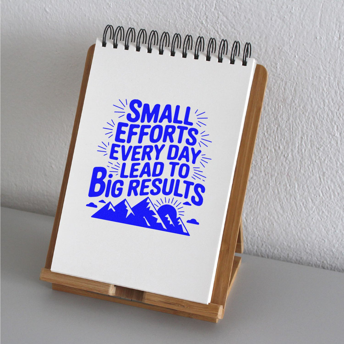 Blue rubber stamp design on a spiral notepad in a wooden stand, reading Small efforts every day lead to big results with mountain graphic.