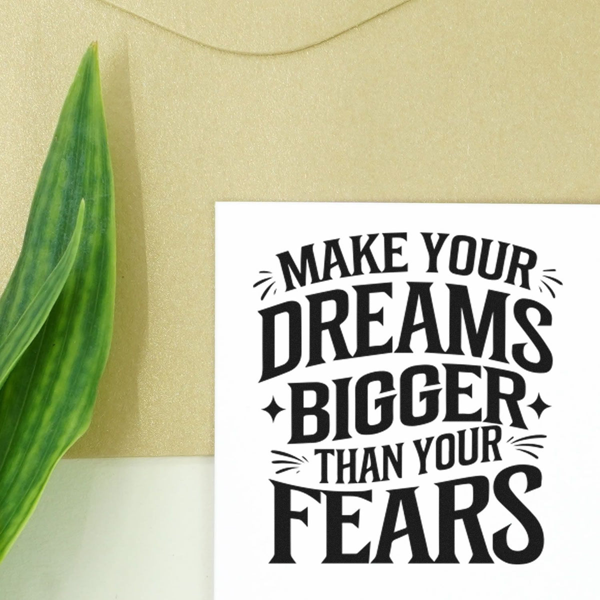 Custom rubber stamp seal with black quote Make your dreams bigger than your fears on white, beside green leaf on gold background.