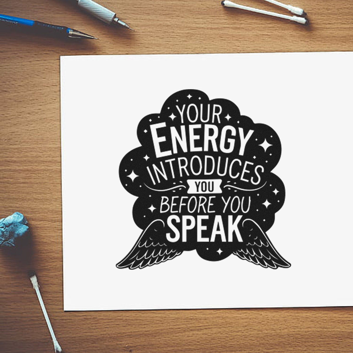 Black custom stamp seal with winged quote on white paper: Your energy introduces you before you speak, on a wooden desk.