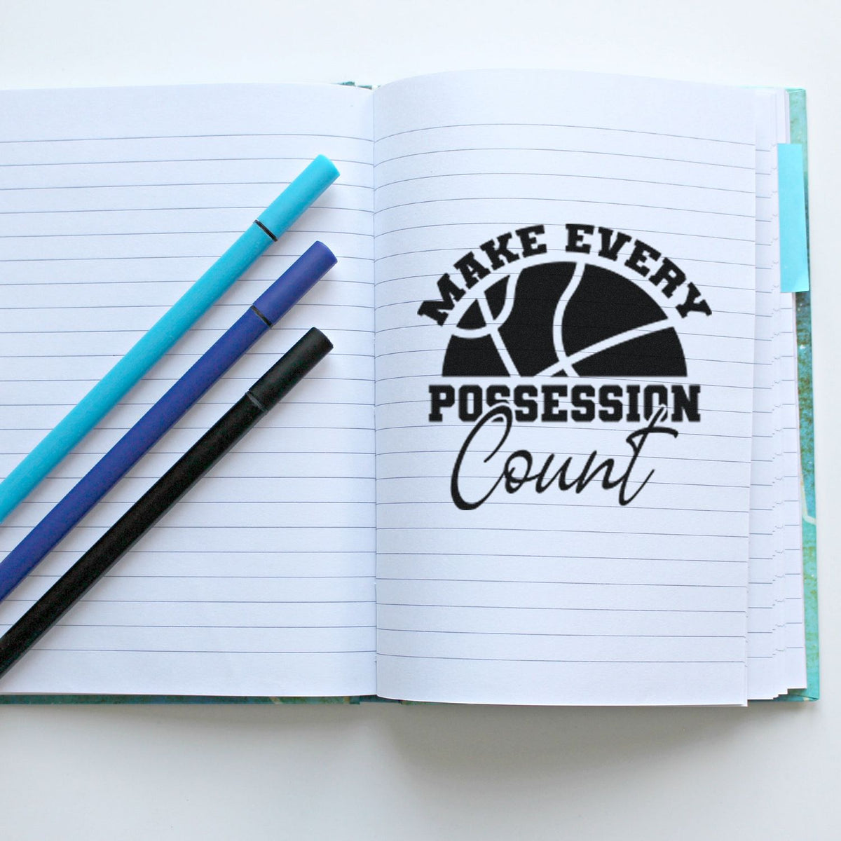 Open lined notebook with black basketball stamp reading Make Every Possession Count and turquoise, purple, and black markers.