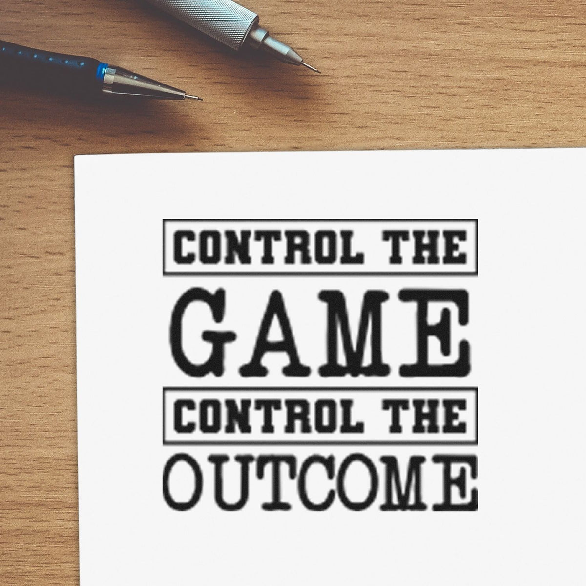 Black stamp and seal design on white paper reading Control the Game, Control the Outcome, with pens on a wood desk.