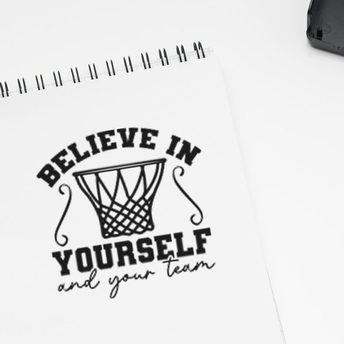 Black rubber stamp impression on white spiral notebook page, with basketball hoop and Believe in yourself and your team.
