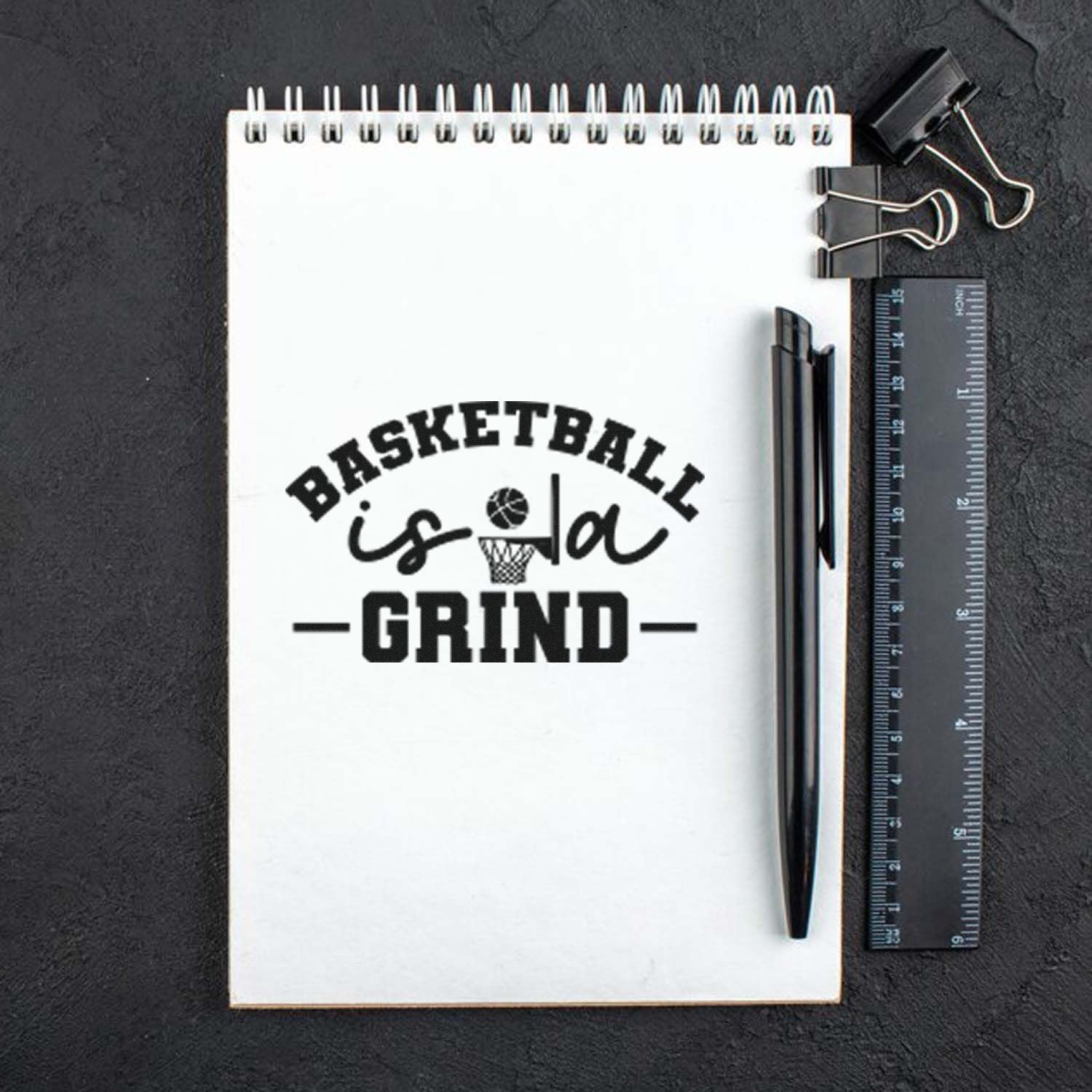 Black basketball-themed stamp on a white spiral notebook, reading Basketball is a Grind, with pen, ruler, and binder clips.