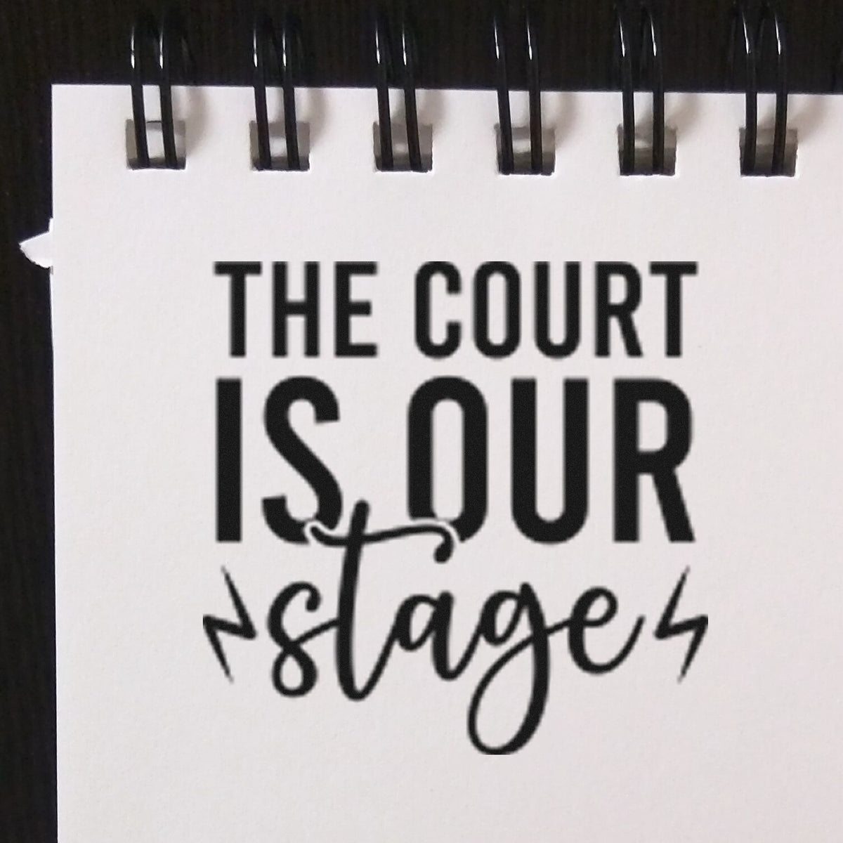 Black-and-white stamp and seal notebook page with spiral binding, reading THE COURT IS OUR stage in bold and script text.