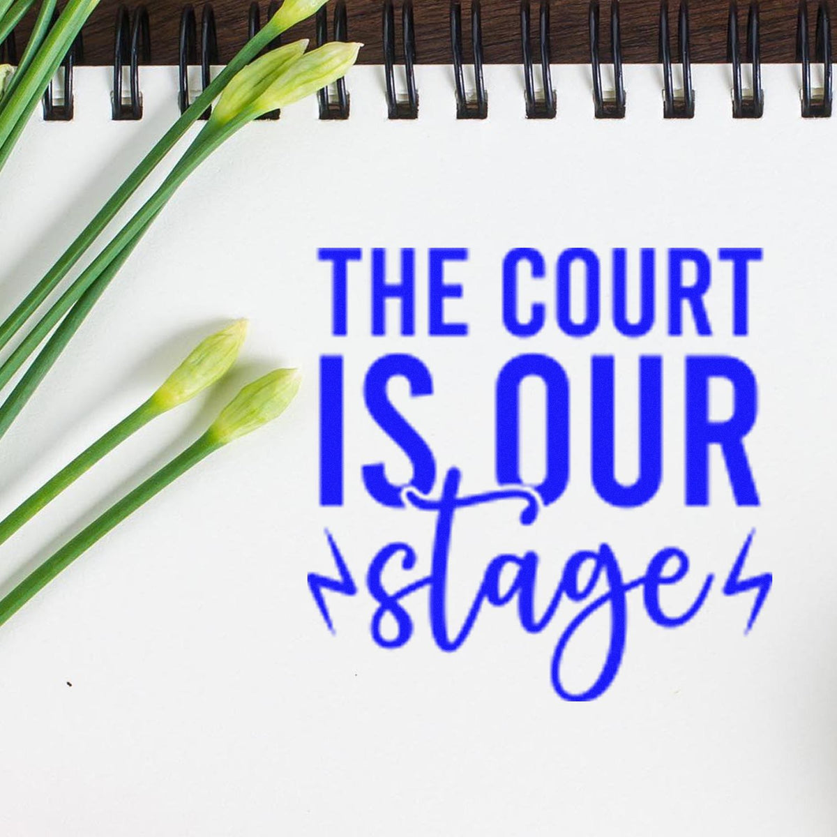Blue stamp and seal text THE COURT IS OUR stage on a white spiral notebook with green flower stems.