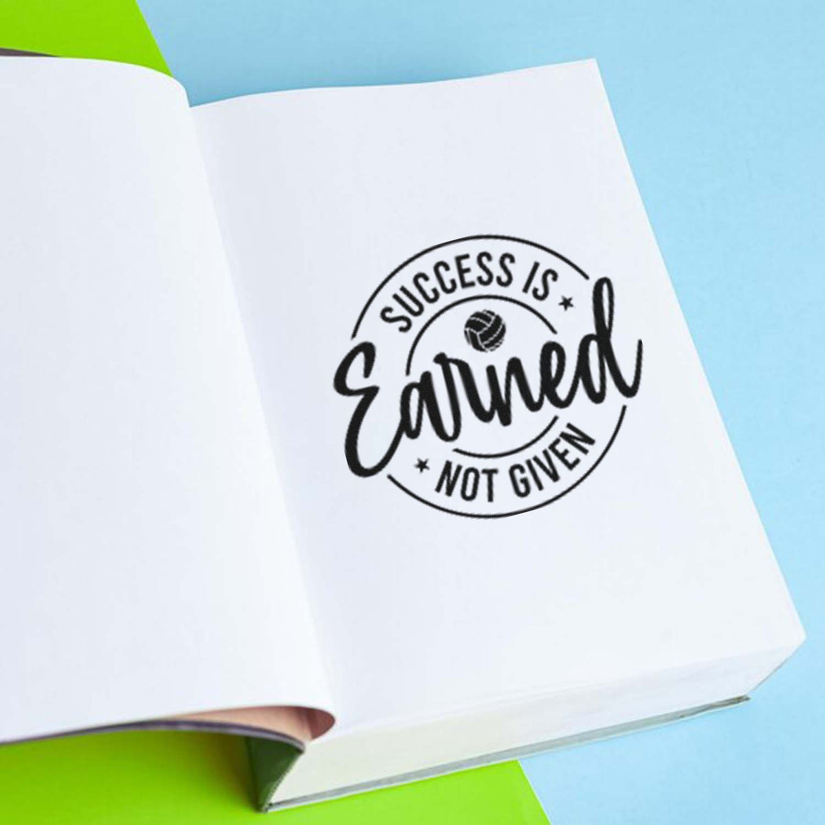 Open white notebook showing a black motivational stamp seal reading Success Is Earned, Not Given on blue and lime green.